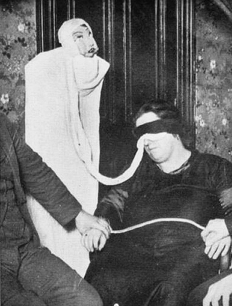 A black and white flash photograph from a 1931 séance showing medium Helen Duncan seated and blindfolded with a dark cloth strip. A thick, white, rope-like strand of "ectoplasm" extends from her nose or mouth, trailing upward into a crude, upright ghostly figure made of a draped white sheet. The figure is topped with a painted papier-mâché mask or doll-like face looking back toward her. A man sitting to the left holds her hand.