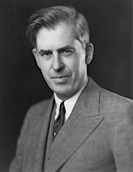 A crisp, black and white studio portrait of American politician Henry A. Wallace, showing him from the chest up against a dark background. He is wearing a structured tweed suit jacket with a matching vest, a white collared shirt, and a dark necktie, looking directly at the camera with a subtle expression.
