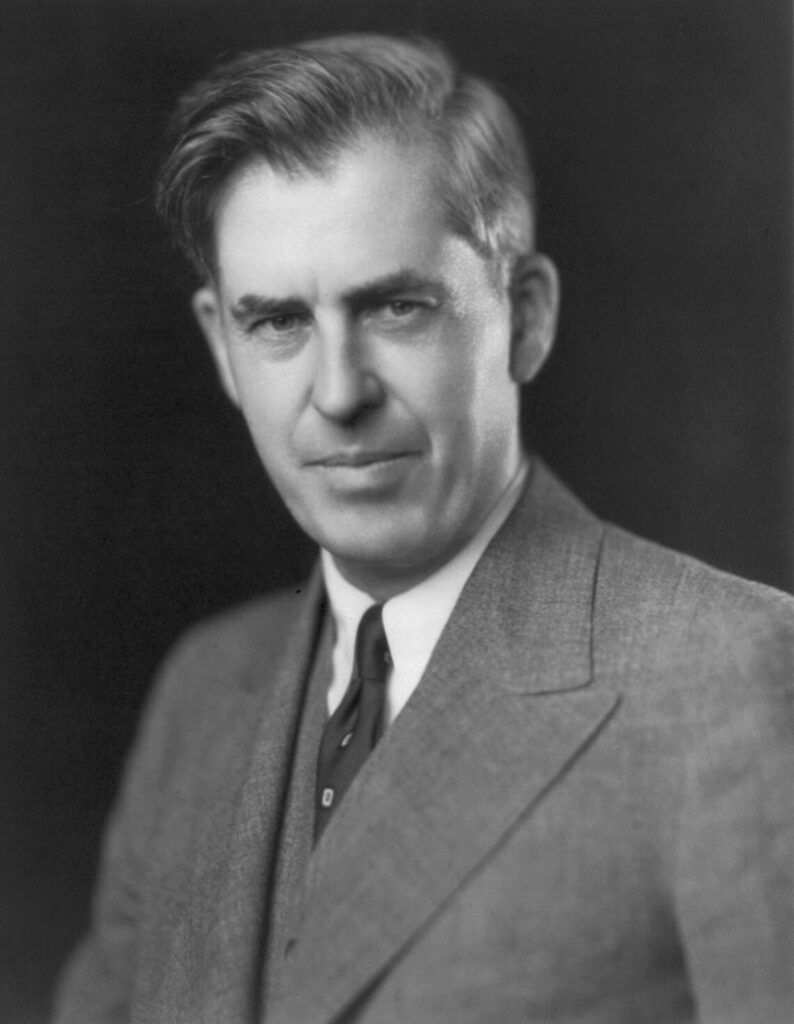 A crisp, black and white studio portrait of American politician Henry A. Wallace, showing him from the chest up against a dark background. He is wearing a structured tweed suit jacket with a matching vest, a white collared shirt, and a dark necktie, looking directly at the camera with a subtle expression.