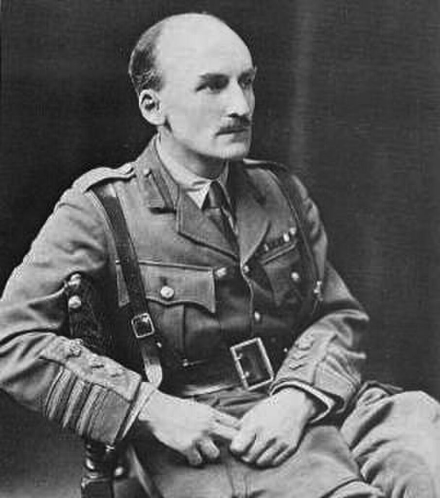 A black and white studio portrait of British Army officer J.F.C. Fuller sitting in a chair, dressed in a military uniform with a Sam Browne belt, mustache, and looking off to the side.
