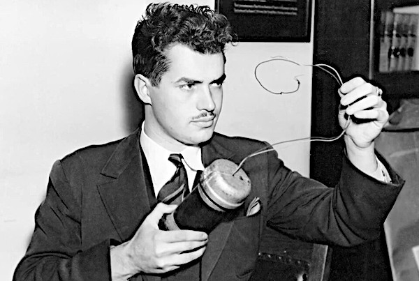 A black and white historical photograph of a young Jack Parsons. He has thick, wavy dark hair, a thin mustache, and wears a dark suit jacket, a white shirt, and a striped necktie. He is holding a cylindrical mechanical device with wires protruding from the top, holding the wire loop up with his left hand as he examines it with a focused gaze
