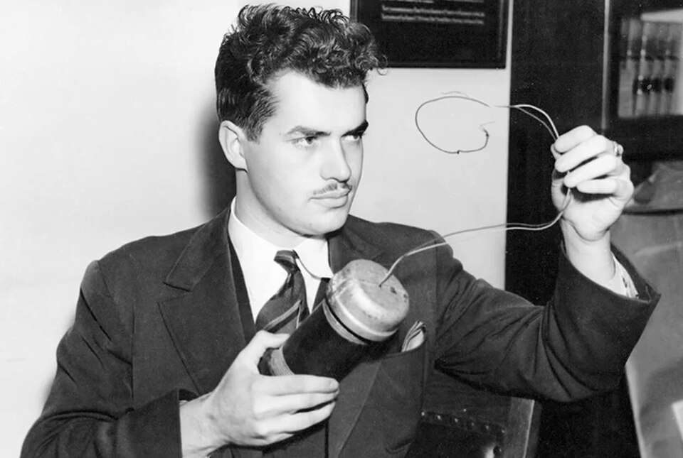 A black and white historical photograph of a young Jack Parsons. He has thick, wavy dark hair, a thin mustache, and wears a dark suit jacket, a white shirt, and a striped necktie. He is holding a cylindrical mechanical device with wires protruding from the top, holding the wire loop up with his left hand as he examines it with a focused gaze