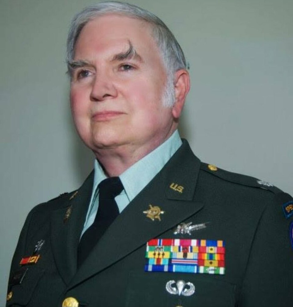 A color portrait of a clean-shaven, older Michael Aquino wearing a dark green U.S. Army dress uniform with a light green collared shirt and black tie. He has short gray hair, prominent sideburns, and a highly distinctive left eyebrow that is long, dark, and dramatically arched upward toward his forehead. His uniform jacket is adorned with gold "U.S." collar insignia, multiple rows of colorful military ribbon bars, and various metallic qualification badges on his chest. He is facing forward and looking slightly off-camera against a plain, light-colored background.