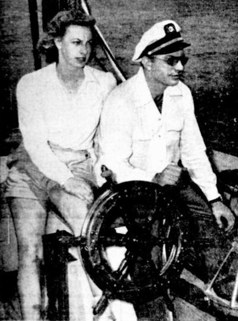 A black and white historical photograph from June 1946 showing a young Sara "Betty" Northrup and L. Ron Hubbard on a boat deck. Hubbard is sitting at a large wooden captain's wheel wearing a white maritime uniform hat, dark sunglasses, and a button-down shirt. Northrup is sitting closely beside him to his left, wearing shorts and a white collared blouse, looking off-camera.