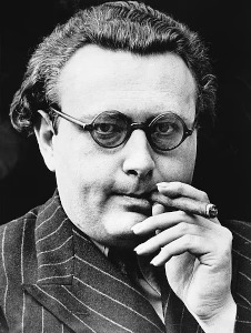 A close-up black and white portrait of astrologer Louis de Wohl. He is facing forward, wearing round, dark-rimmed spectacles and a pinstriped suit jacket. His right hand is raised near his face, holding a burning cigar between his fingers, and he has a focused, intense gaze directed into the camera.
