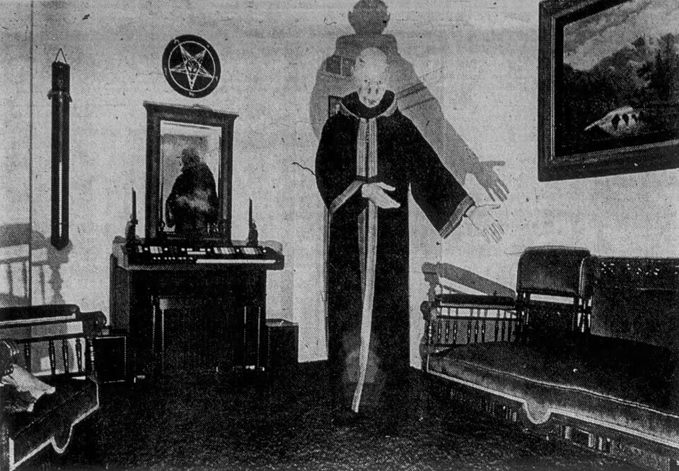 A grain-textured, black and white historical photograph from inside a darkly decorated ritual room. In the center stands U.S. Army psychological operations officer and occultist Michael Aquino. He is wearing long, dark ceremonial robes with light-colored trim and has his left arm extended outward. Behind him, a stark light projects a large, looming shadow of his silhouette onto the wall. The room features a dark couch, a framed painting, an electronic organ console beneath a vertical wall mirror, and a circular wall plaque displaying an inverted pentagram.