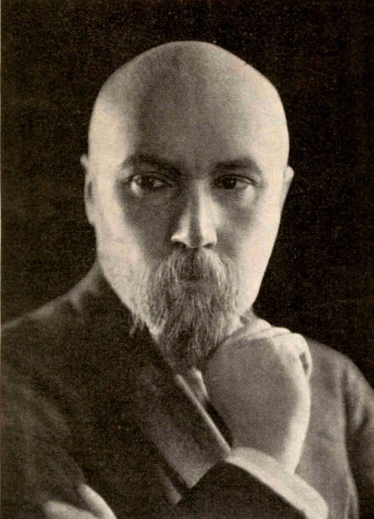 A sepia-toned vintage portrait photograph of Nicholas Roerich. The subject is a bald man with a prominent beard and mustache, resting his chin thoughtfully on his hand while staring directly forward into the camera with an intense expression.