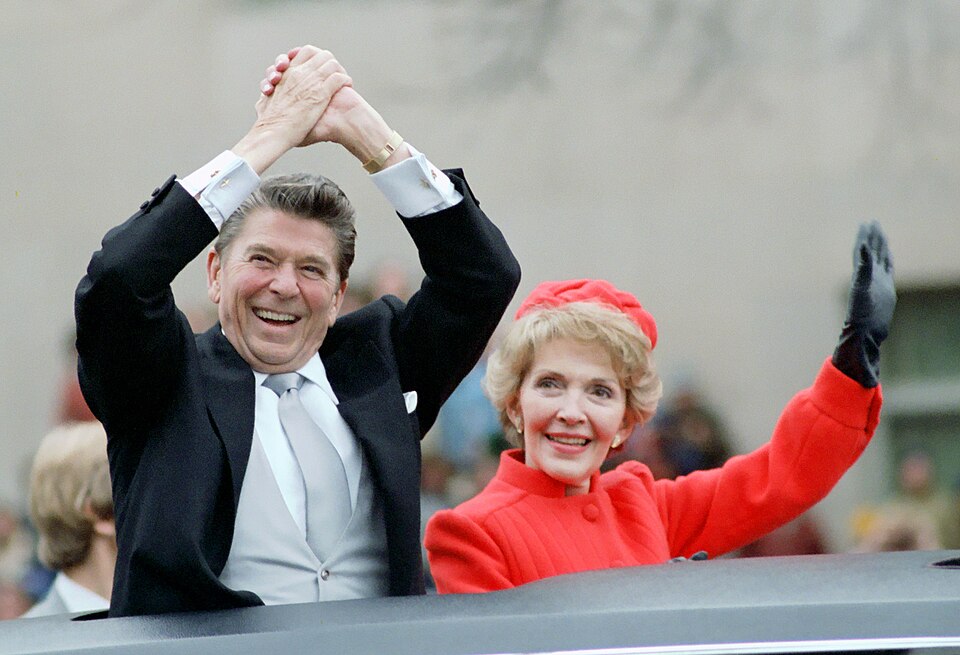 A color photograph of President Ronald Reagan and First Lady Nancy Reagan standing up in an open-top vehicle during a public parade. On the left, Ronald Reagan is laughing joyfully with his hands clasped together over his head in a victory wave, wearing a black formal suit coat with a light gray vest and a matching silk tie. On the right, Nancy Reagan is smiling and waving a gloved left hand toward the crowd, dressed in a bright red pillbox hat and a matching high-collared red wool coat.