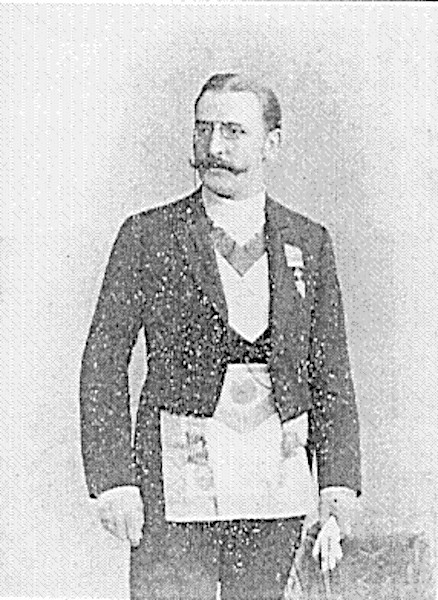 A grain-textured, black and white historical photograph of German occultist Theodor Reuss. He is standing and facing forward while looking slightly to the side, sporting a prominent mustache and round spectacles. He is dressed in formal Masonic evening wear, complete with a dark tailcoat, white collared shirt, decorative neck regalia, and a large, rectangular Masonic apron tied around his waist.