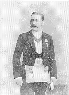 A grain-textured, black and white historical photograph of German occultist Theodor Reuss. He is standing and facing forward while looking slightly to the side, sporting a prominent mustache and round spectacles. He is dressed in formal Masonic evening wear, complete with a dark tailcoat, white collared shirt, decorative neck regalia, and a large, rectangular Masonic apron tied around his waist.