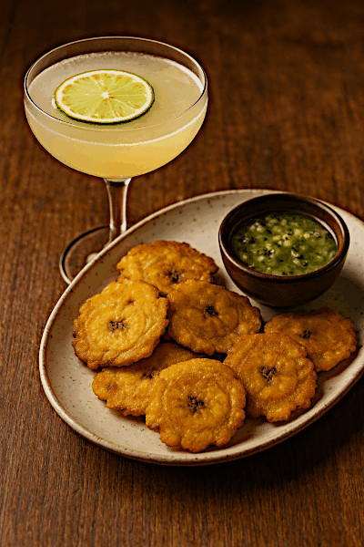 A Hemingway Daiquiri cocktail in a coupe glass with a lime wheel garnish, served beside a plate of crispy golden tostones with a small bowl of green mojo sauce on a wooden table.