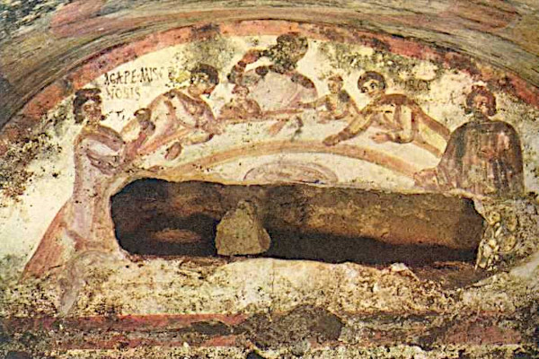 An ancient, faded fresco painted on the arched ceiling of a stone catacomb tomb. It depicts a group of early Christians gathered around a semi-circular table for a communal meal. Seven figures are visible, gesturing toward one another and sharing food. On the left side of the scene, Latin text reading "AGAPE MISCE NOBIS" is painted on the wall. The lower portion of the image shows an open, horizontal tomb carved directly into the rock beneath the painting.