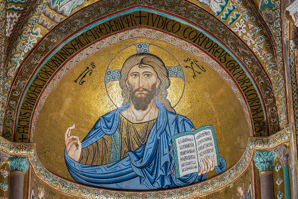 A majestic, gold-leaf Byzantine mosaic filling the semi-dome of a cathedral apse. In the center, a monumental half-length depiction of a bearded Jesus stares forward with a stern expression. He wears a blue mantle over a gold tunic and holds a large open book with inscriptions written in both Greek and Latin scripts. His right hand is raised in a traditional gesture of blessing. A large, jeweled cross-halo surrounds his head against a solid shimmering gold background, bordered by an arch inscribed with prominent Latin lettering.
