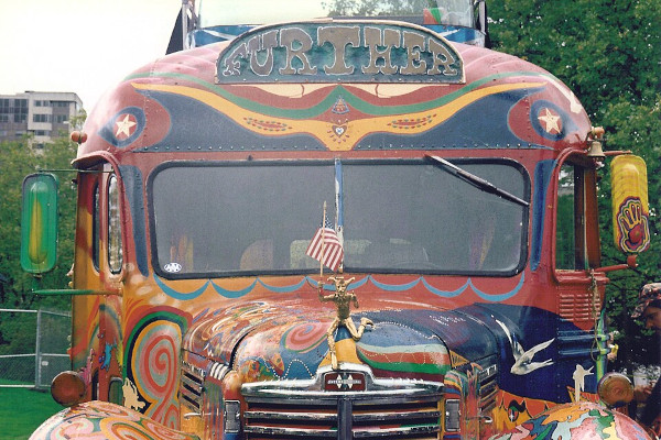 The front of the vintage "Furthur" bus. It is covered in vibrant, hand-painted psychedelic swirls. A prominent sign above the windshield reads "FURTHUR". A small jester figure acts as a hood ornament. This jester holds a miniature American flag. The side mirrors feature bright, hand-painted designs. One mirror displays a colorful handprint graphic.