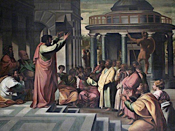 A large Renaissance oil painting depicting a grand public square in ancient Athens. On the left, Saint Paul stands elevated on stone steps with both arms raised high in a dramatic preaching gesture. He wears a flowing pink mantle over a green robe. A diverse crowd of bearded Greek philosophers, citizens, and elite figures surround the steps, listening intently with varied expressions of skepticism, contemplation, and curiosity. In the background stands a prominent circular temple with classical columns, topped by a large bronze statue of a Roman soldier holding a spear and shield.