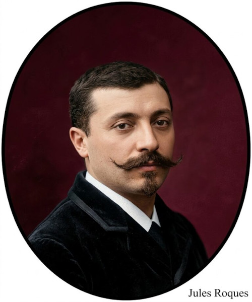 Colorized, oval-framed portrait of Jules Roques, featuring his distinctive upturned waxed mustache, soul patch, and dark velvet jacket against a deep red background.