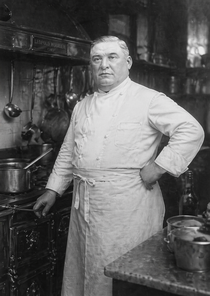 Black and white portrait of Chef Leopold Mourier in a professional kitchen, wearing a traditional double-breasted white chef's coat and apron.
