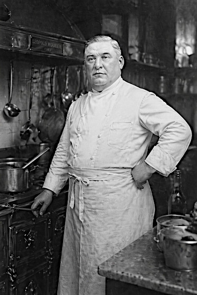 Black and white portrait of Chef Leopold Mourier in a professional kitchen, wearing a traditional double-breasted white chef's coat and apron.