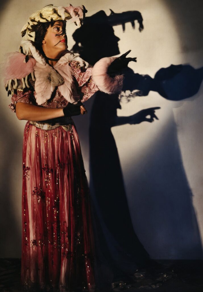 A colorized photograph of the performer O’dett in elaborate drag, wearing a ruffled pink gown and a large feathered hat, while casting a dramatic, distorted shadow against a plain wall.