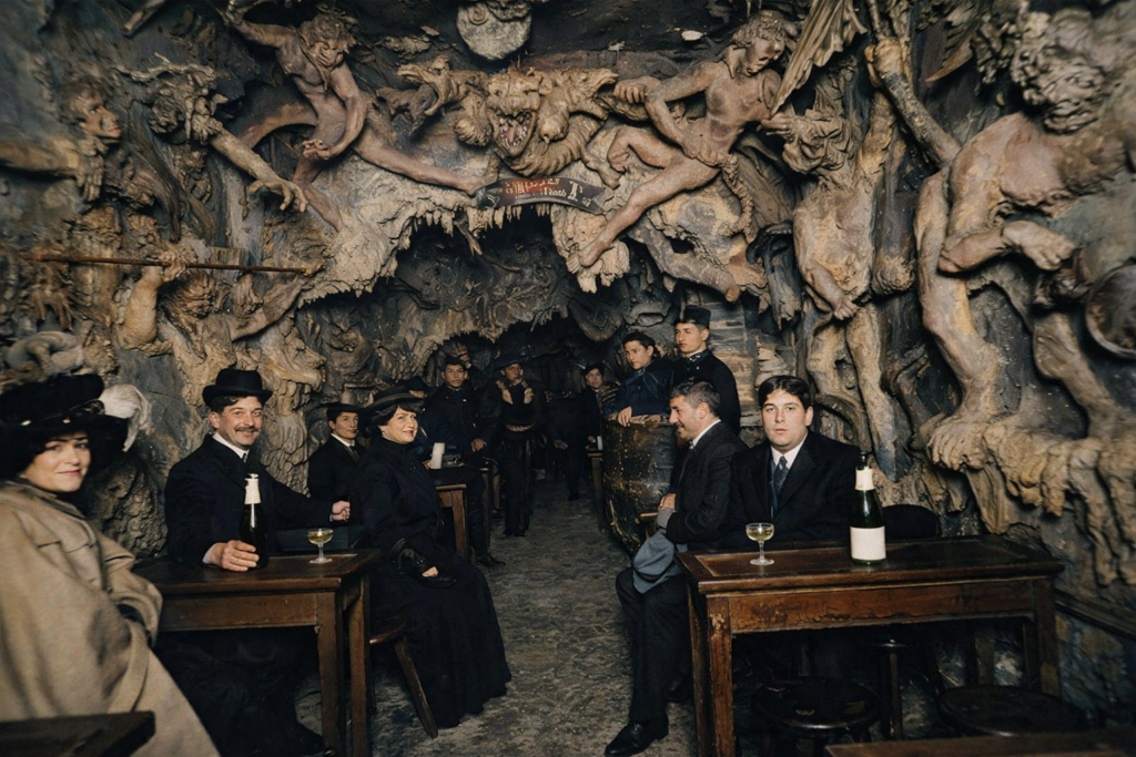 Colorized vintage photo of patrons drinking inside the Cabaret de l'Enfer in Paris, surrounded by plaster sculptures of demons and grotesque figures.
