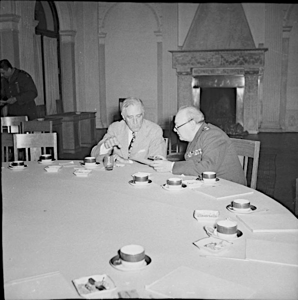 A grainy, high-contrast vintage photograph capturing a moment of quiet exhaustion after a summit. Two elderly men—Winston Churchill in a dark suit and Franklin D. Roosevelt in a gray suit—sit close together at a massive, circular conference table. The vast table surface is messy, scattered with used papers and empty white coffee cups. All surrounding chairs are pushed back and completely empty, with the two leaders appearing isolated in a deep, private discussion under soft indoor lighting.