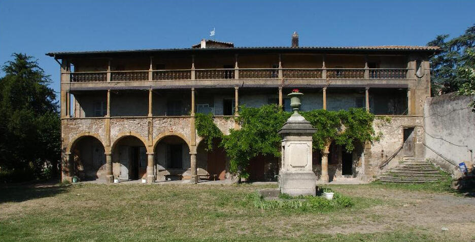 A wide, eye-level outdoor photograph of the historic Villa du Perron under a clear blue sky. The building features a multi-tiered facade with an open stone loggia on the ground floor consisting of several rounded arches supported by columns. Above the arches run two levels of covered wooden balconies with decorative balustrades, supported by slender pillars. Lush green climbing vines cover a portion of the central ground floor walls. In the foreground is a grassy, unpaved courtyard featuring a central stone pedestal monument topped with an ornamental urn, and a curved stone staircase leads to an upper doorway on the far right.
