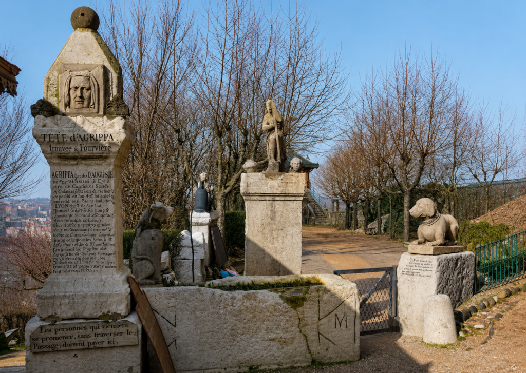 A color photograph of the historic ruins along the Passage Gay path on the Fourvière hillside under a clear blue sky. On the left stands a large, multi-tiered stone pedestal monument featuring a carved stone portrait bust labelled "TETE D'AGRIPPA Trouvée à Fourvière" over a long carved block of French text. In the center, a narrow stone pillar is topped with a full-body classical statue, while a lower stone column to its left features a small metal urn. On the far right, a stone statue of a dog sits atop a block next to an open metal gate. The unpaved path is bordered by bare trees and leads toward a distant iron fence overlooking the city rooftops of Lyon in the background.