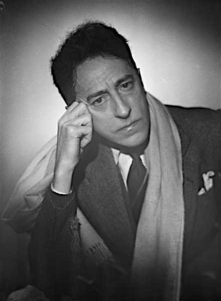 A black and white studio photograph of Jean Cocteau as an older man, resting his head thoughtfully on his right hand. He is wearing a dark tweed suit jacket, a white collared shirt with a dark tie, and a thick, light-colored woolen scarf draped loosely around his neck against a minimalist background.