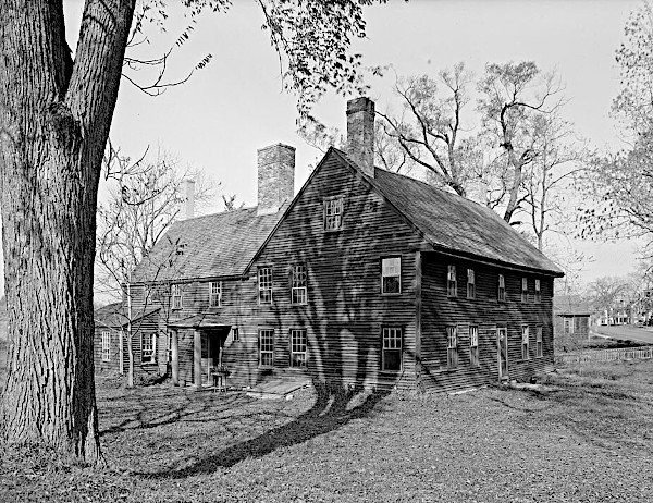 An archival black and white photograph of an early 18th-century, two-story colonial wooden saltbox house with multiple brick chimneys, surrounded by bare trees casting long shadows across the grass and facade.