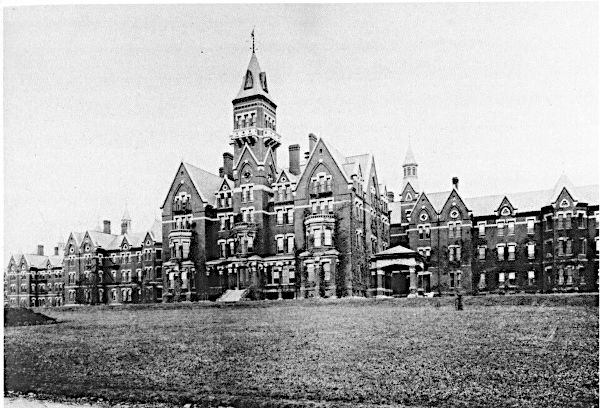 An archival black and white photograph from the 1890s showing the massive Victorian Gothic brick facade of Danvers State Hospital, featuring a soaring central clock tower and sprawling administrative wings fronted by a wide, empty grass lawn.