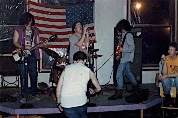 A full band live performance photograph captures GNP (Grossest National Product) playing on a low, triangular wooden stage. A large American flag hangs on the wall directly behind the band as a backdrop. From left to right on stage: Chris Hendrix plays a sticker-covered electric guitar while wearing a purple sleeveless shirt over a graphic tee and blue jeans; Spike stands center-stage singing directly into a microphone; a drummer is partially visible behind a compact drum kit; and a bassist with long shaggy hair facing away from the audience plays an electric bass guitar. In front of the stage, a fan in a sleeveless white shirt moshes with their back to the camera. On the far right, another person in a yellow t-shirt sits on a chair, watching the band intently.