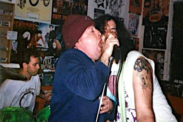 A grainy, candid color photograph captures a high-energy performance by the Birmingham, Alabama punk band GNP (Grossest National Product) inside a crowded, poster-lined room. In the foreground, two male vocalists lean heavily into a single microphone. The vocalist on the left wears a brown beanie and a blue long-sleeve shirt, his mouth wide open as he screams aggressively into the mic. The vocalist on the right has long, dark, disheveled hair, a prominent arm tattoo, and a sleeveless white shirt, singing passionately with his eyes closed. In the background on the left, the drummer sits behind a green drum kit, watching the performance. The wall behind them is plastered with underground music flyers, band gig posters, and a sign reading "New Arrivals."
