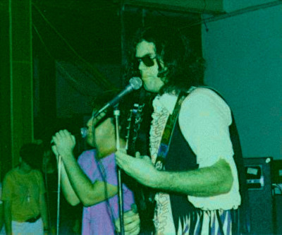 A grainy color photograph with a heavy green tint captures a live performance by Chris and Spike from the Birmingham punk band GNP (Grossest National Product) at The Loft. In the foreground, Spike stands on the right, wearing sunglasses, a collared shirt, and a dark vest, singing into a microphone while holding an electric guitar. To his left, Chris leans forward into his own microphone stand, gripped with both hands, wearing a purple shirt as he delivers vocals. In the dim background on the far left, another individual is partially visible watching the performance.