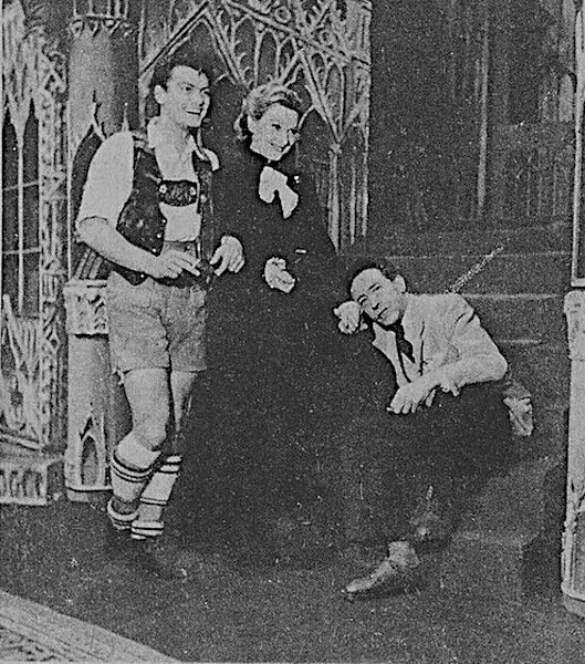 An archival black and white photograph from a 1946 theatrical production, showing actor Jean Marais standing on the left in a traditional embroidered vest, shorts, and long striped socks, standing next to actress Edwige Feuillère in a long dark gown, while Jean Cocteau sits smiling on a set of wooden stairs to the right, leaning his head on his hand.