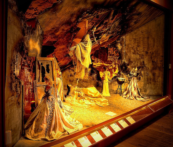 A museum diorama display case showing a dramatic theatrical stage set filled with mannequins wearing ornate, historical ball gowns. The setting features a surreal, fractured environment with crumbling stone walls, a broken glass door, an overhead rocky canopy, and golden warm lighting illuminating a figure reclining on a bed in the center.