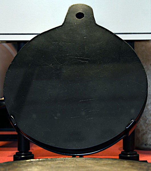 A close-up photograph of a circular, polished black volcanic glass mirror with a rounded handle tab at the top featuring a single pierced hole, resting on a museum display stand against a neutral background.