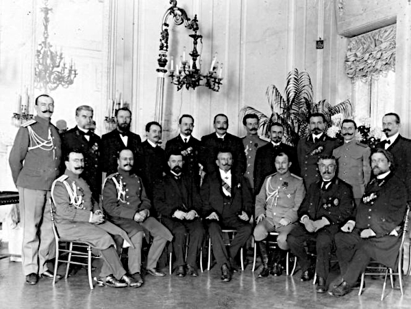An archival black and white photograph from 1905 featuring fifteen stern men of the Tsarist Russian secret police posing together indoors beneath an ornate chandelier, with several officers in formal military uniforms with buttons and white chest cords alongside investigators in dark civilian suits.