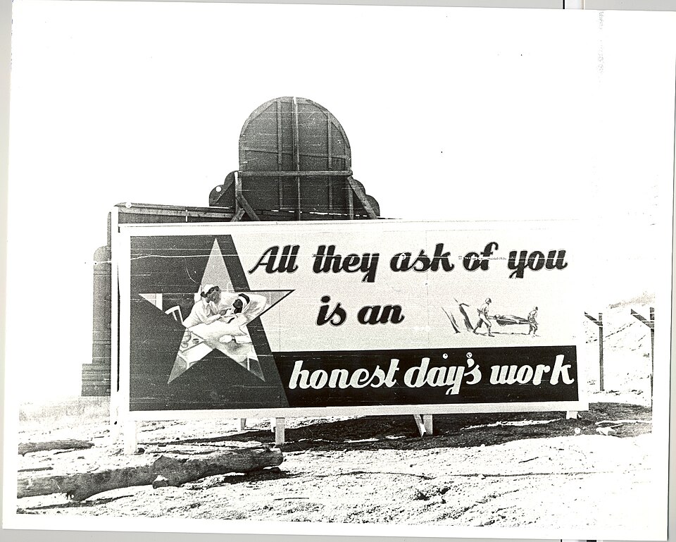 A black and white historical photograph of a large outdoor billboard standing in a cleared dirt and sand area with logs on the ground. The sign features a large star graphic containing an illustration of a woman working at a machine, alongside text that reads "All they ask of you is an honest day's work." On the right side of the billboard is a small illustration of workers carrying material.
