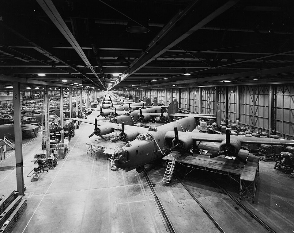 A dramatic black and white historical photograph capturing a massive, seemingly endless industrial assembly line inside an aircraft factory hangar. Rows of heavy four-engine B-24 Liberator bombers are positioned wing-to-wing down a long production bay under a dense network of structural steel roof trusses, overhead cranes, and industrial factory lighting.