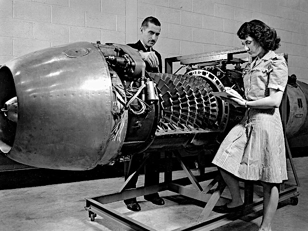 A vintage black and white documentary photograph from 1946 showing a male engineer and a female technician examining a large, metallic aircraft turbojet engine on a rolling steel assembly stand. The technician on the right stands in a striped dress writing notes on a clipboard, while the engineer stands behind the exposed, multi-stage internal compressor blades and turbine rows.