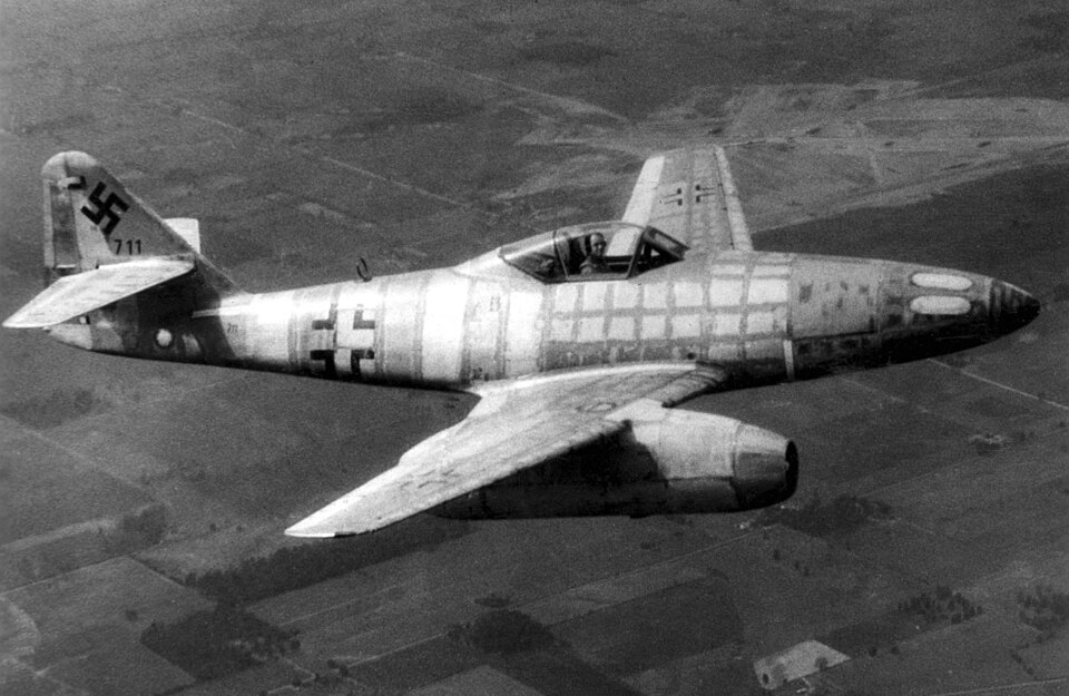 An archival black and white photograph of a Messerschmitt Me 262 jet fighter in mid-flight over a landscape of rural fields. The aircraft features Luftwaffe military cross insignia on the fuselage, a swastika on the tail fin, and distinctive underslung jet engine pods beneath its swept wings.