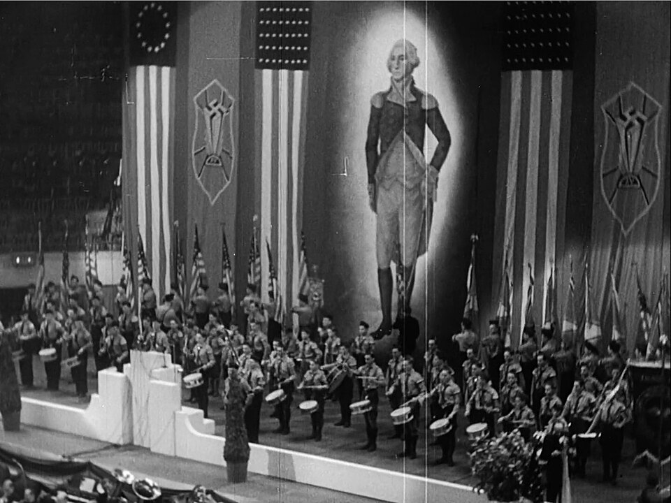 An archival black and white photograph from the 1939 German American Bund rally inside Madison Square Garden, showing a massive stage lined with long American flag banners flanking a colossal central portrait painting of George Washington wrapped in a glowing white halo. On either side of the portrait hang vertical banners emblazoned with a modified swastika insignia, while rows of uniformed men playing marching snare drums line the multi-tiered white stage below.