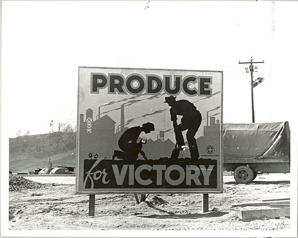 A black and white historical photograph of a large outdoor billboard standing in a dirt field. The sign reads "PRODUCE for VICTORY" in bold letters and features a graphic silhouette illustration of a man shoveling and a person kneeling to garden against an industrial background of smoking factory smokestacks. A covered military transport truck is parked on the right.