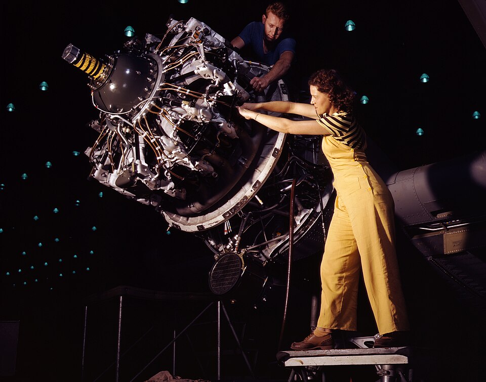 A vibrant vintage color photograph showing a female factory worker in yellow overalls standing on a platform to assemble a massive aircraft engine. A male worker stands behind the exposed machinery, working beneath a dark ceiling dotted with patterns of green indicator lights.